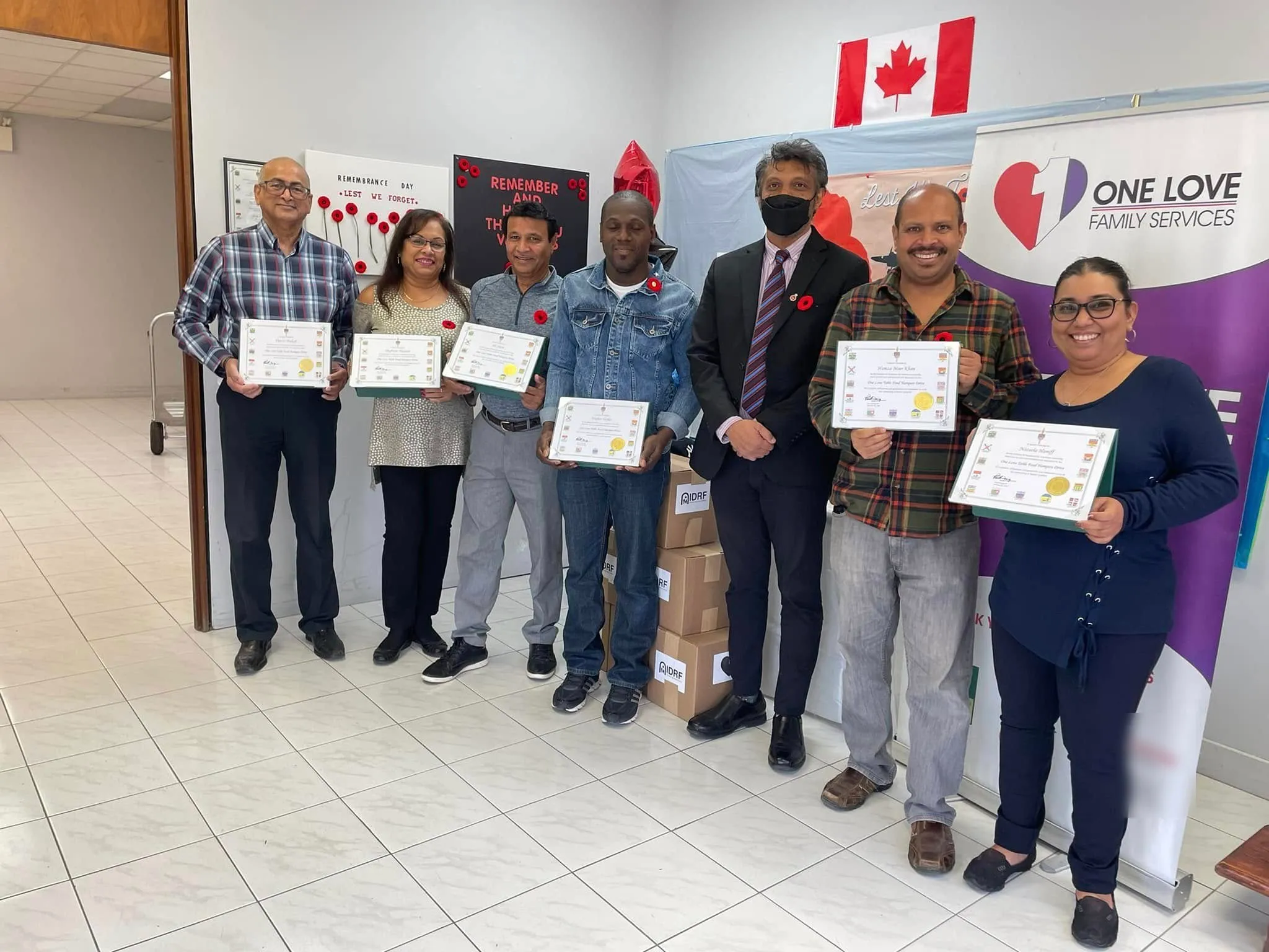 One Love volunteers holding certificates of appreciation for the One Love Table Food Hamper Drive, beside a One Love Family Services banner